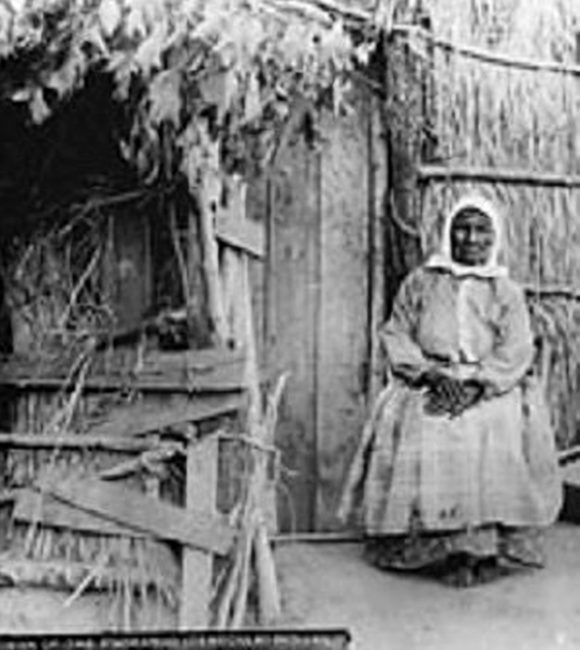 A Payómkawichum woman standing outside a traditional structure, early photograph